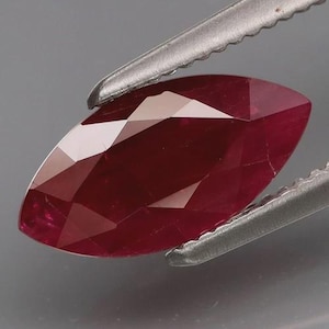 Pode incluir: Gema de rubi com corte marquesa, segurada por pinças prateadas. O rubi tem uma cor vermelha profunda com múltiplas facetas que refletem a luz. A gema está sobre um fundo cinza.