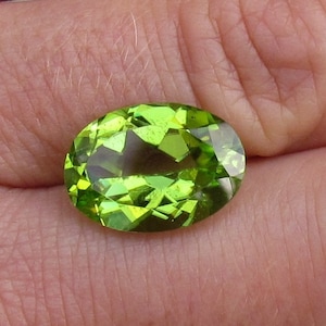 May include: A faceted, oval-cut peridot gemstone. The gem is a vibrant, translucent green color, with multiple facets that reflect light. The peridot is held in a hand, showcasing its clarity and brilliance. This gemstone is ideal for jewelry.