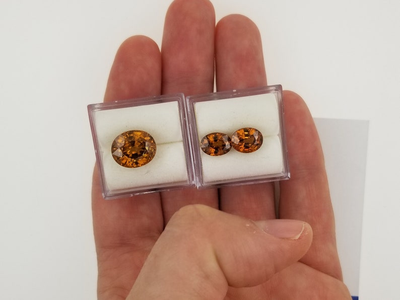 May include: Two oval-shaped gemstones in clear plastic boxes. The gemstones are a deep orange color with a slight reddish hue.