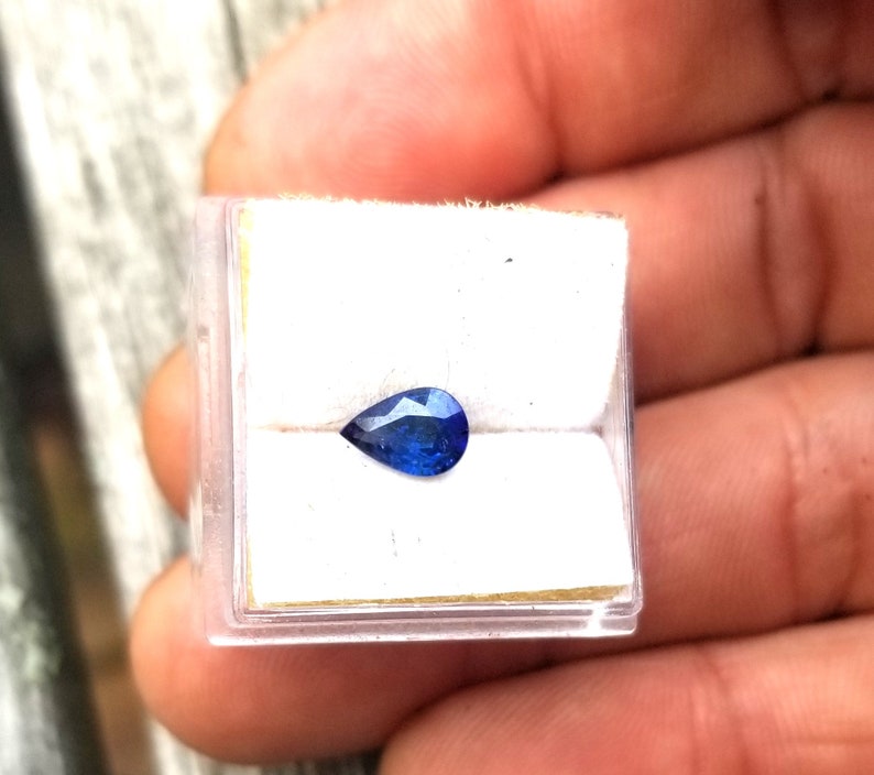 Puede incluir: Una piedra preciosa de zafiro azul en forma de pera en una peque&ntilde;a caja blanca.