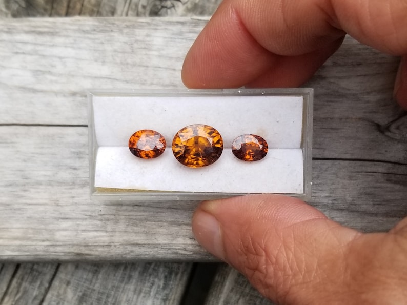 May include: Three oval-shaped gemstones in a white display box. The gemstones are orange and brown with a slight sparkle. The gemstones are arranged in a row from largest to smallest.