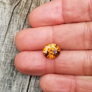 May include: A close-up of an oval-shaped, orange gemstone with a faceted cut. The gemstone is held in a hand, and the background is a wooden surface.