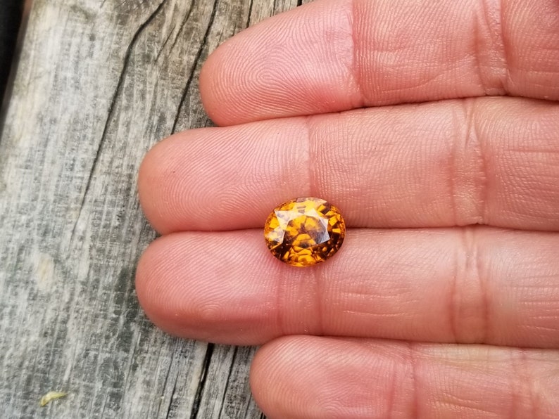 May include: A close-up of an oval-shaped, orange gemstone with a faceted cut. The gemstone is held in a hand, and the background is a wooden surface.