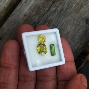May include: A small white box with three gemstones inside. Two of the gemstones are yellow and oval shaped. The third gemstone is green and rectangular.