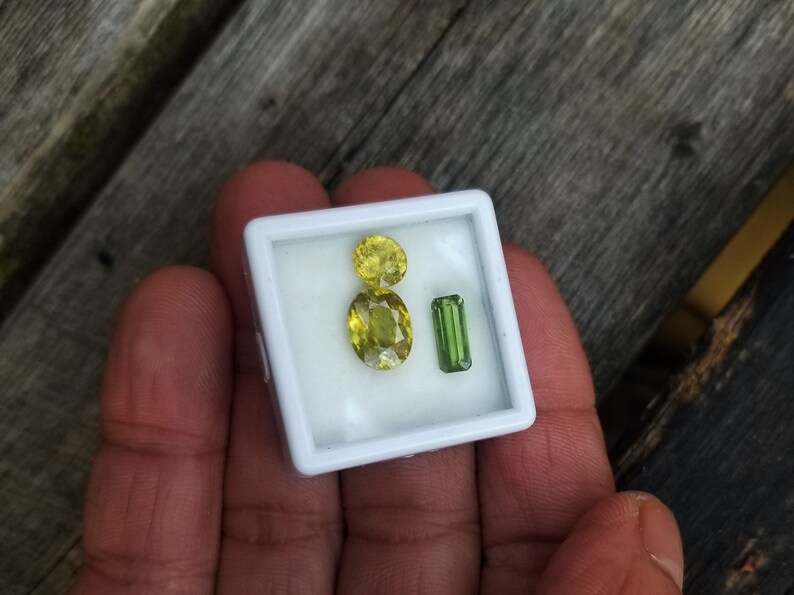 May include: A small white box with three gemstones inside. Two of the gemstones are yellow and oval shaped. The third gemstone is green and rectangular.