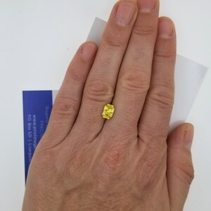 K&ouml;nnte beinhalten: Ein leuchtend gelber, kissenf&ouml;rmig geschliffener Edelstein, gehalten in einer Hand vor wei&szlig;em Hintergrund. Die Facetten des Steins fangen das Licht ein und zeigen seinen Glanz. Eine kleine blaue und wei&szlig;e Karte ist teilweise sichtbar.