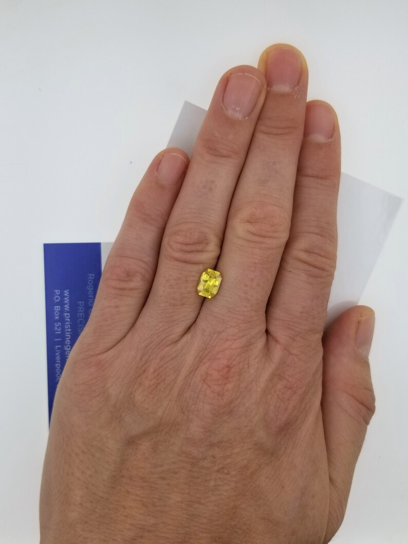 K&ouml;nnte beinhalten: Ein leuchtend gelber, kissenf&ouml;rmig geschliffener Edelstein, gehalten in einer Hand vor wei&szlig;em Hintergrund. Die Facetten des Steins fangen das Licht ein und zeigen seinen Glanz. Eine kleine blaue und wei&szlig;e Karte ist teilweise sichtbar.