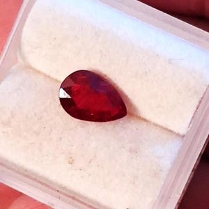 May include: A pear-shaped, faceted, deep red gemstone, possibly a ruby, rests on a white, textured surface within a clear plastic case. The gem's cut allows light to reflect, creating a brilliant appearance. The case is held by a person.