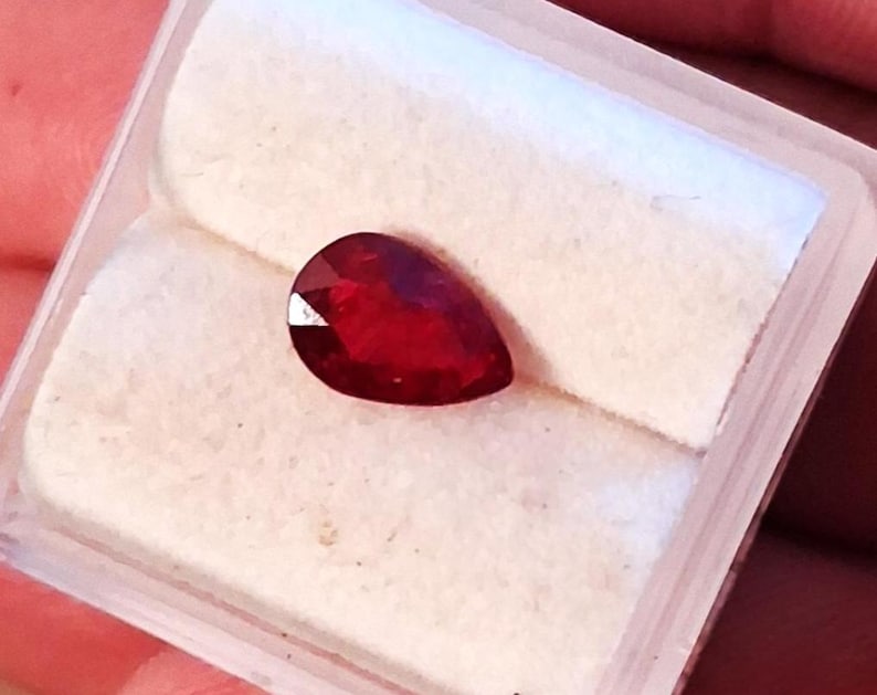 May include: A pear-shaped, faceted, deep red gemstone, possibly a ruby, rests on a white, textured surface within a clear plastic case. The gem's cut allows light to reflect, creating a brilliant appearance. The case is held by a person.