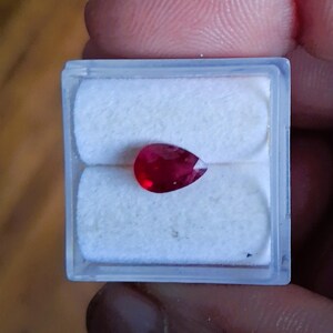 May include: A pear-shaped, deep red gemstone, possibly a ruby, is displayed in a clear plastic case with a white, cushioned interior. The gem's facets catch the light, showcasing its rich color and clarity. The case is held by a person's fingers.