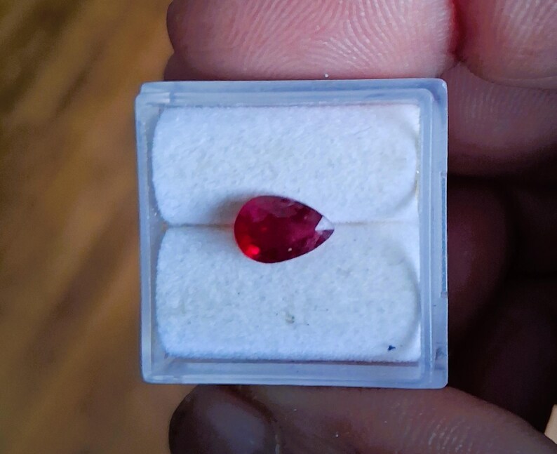 May include: A pear-shaped, deep red gemstone, possibly a ruby, is displayed in a clear plastic case with a white, cushioned interior. The gem's facets catch the light, showcasing its rich color and clarity. The case is held by a person's fingers.