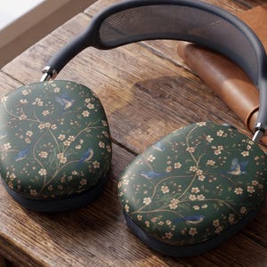 May include: Over-ear headphones featuring a dark green exterior shell decorated with a repeating pattern of small light pink flowers and blue birds. The headband includes a dark gray mesh top section and adjustable metal arms connecting to the ear cups.
