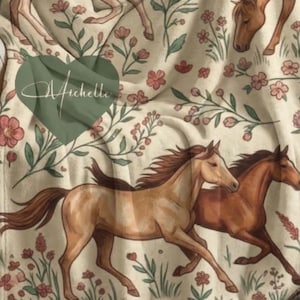 Könnte beinhalten: Cremefarbene Decke mit einem Pferde- und Blumendesign. Die Decke hat ein grünes Herz mit dem Aufdruck "Arichelle". Die Decke ist über einer Person drapiert, die in einem Stuhl sitzt.