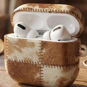 May include: A brown and white cowhide AirPods Pro case with a stitched design. The case is open, revealing white AirPods. A leather keychain and keys are in the foreground. The case has a small handle.
