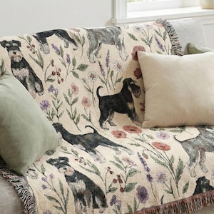 Op de afbeelding: Een beige sprei met een herhalend patroon van Schnauzer honden en bloemmotieven, waaronder lavendel en klaprozen. De sprei, met franjes, is gedrapeerd over een grijze bank met kussens.