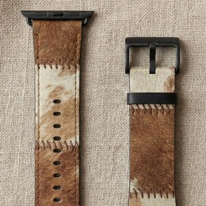以下が含まれることがあります： 2つの時計バンドセット。バンドは茶色とクリーム色の牛革製で、黒い金具が付いています。革はコントラストのある糸で縫い合わされています。スマートウォッチに合うように設計された時計バンドです。
