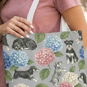 May include: A gray tote bag featuring a pattern of Schnauzer dogs and colorful hydrangea flowers in shades of blue, pink, and white. The bag has a white strap and is held by a person wearing a pink t-shirt.