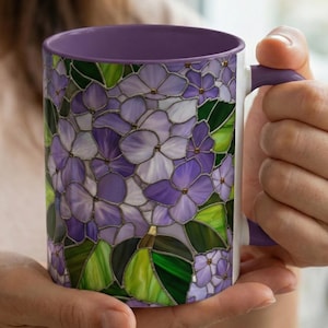 May include: A white ceramic mug with a purple interior and handle. The exterior features a stained-glass design of purple and white hydrangea flowers with green leaves. The mug is held by a person.