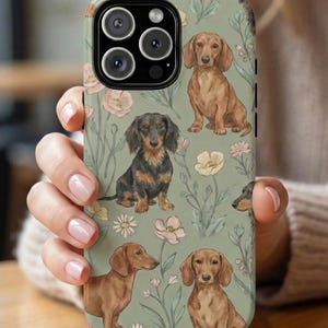 May include: A phone case featuring a repeating pattern of dachshund dogs in various poses and colors, set against a muted green background with floral accents. The case has a camera cutout and black edges.