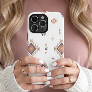 Puede incluir: Funda de teléfono blanca con un patrón geométrico en tonos marrón, azul y dorado. La funda tiene un recorte para la cámara y la sostiene una persona que lleva una camisa rosa y una rebeca color crema.