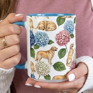 May include: White ceramic mug with a blue handle and interior. The mug features a pattern of golden retrievers and colorful hydrangea flowers. The person holding the mug is wearing a pink shirt and a white cardigan.