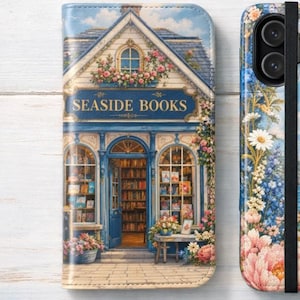 May include: A folding phone case featuring a printed illustration of a bookstore storefront labeled SEASIDE BOOKS on the left, and a coastal scene with a sailboat on the water, flowers, and a black elastic strap on the right side.