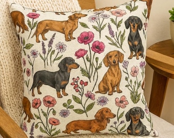 May include: A decorative square pillow featuring a repeating pattern of dachshund dogs in various poses, interspersed with pink and purple flowers and green foliage on a cream background. The pillow is placed on a wooden chair with a cream-colored knitted throw.