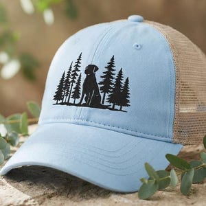 Può includere: Cappellino da baseball azzurro con retro in rete beige. La parte anteriore del cappellino presenta una silhouette nera di un cane seduto davanti a una foresta di pini. Il cappellino ha una visiera curva e un cinturino regolabile.