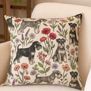 May include: Square cushion with a cream background featuring a repeating pattern of Schnauzer dog illustrations, red poppies, and other floral elements. The cushion is placed on a cream-coloured armchair.