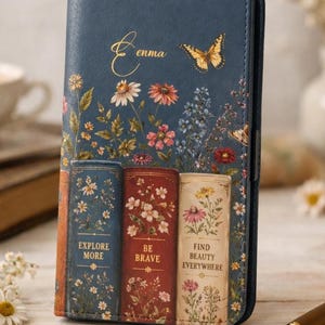 May include: A dark blue rectangular phone case featuring a printed design of three vintage books with floral patterns. The text reads "Enma", "EXPLORE MORE", "BE BRAVE", and "FIND BEAUTY EVERYWHERE". A yellow butterfly is printed above the books.