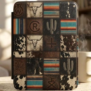 May include: A leather tablet case with a patchwork design featuring a steer skull, cactus, and striped patterns in brown, black, and turquoise. The case has a black spine and is sitting on a wooden surface.