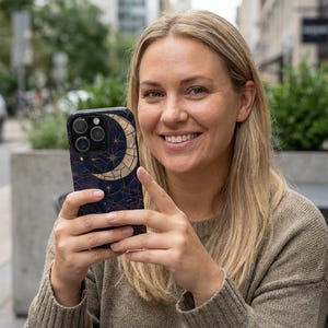 以下が含まれることがあります： ネイビーブルーのスマートフォンケース。金色の三日月と星のデザイン。ケースは光沢があり、人が持っています。