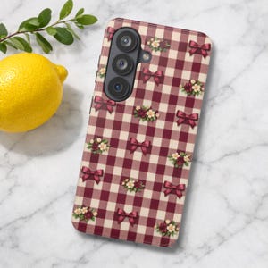 May include: A rectangular smartphone case featuring a maroon and cream gingham check pattern. The design is decorated with small, repeating illustrations of dark red bows and clusters of tiny white and red flowers scattered across the surface.