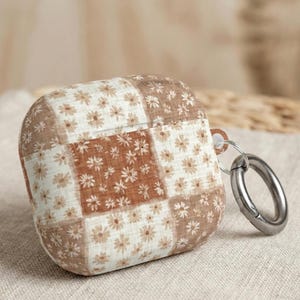 May include: A brown and white floral patterned AirPod case with a silver carabiner clip. The case features a patchwork design with small daisy-like flowers. The background is a neutral beige colour.