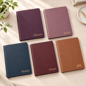 May include: Five rectangular notebooks with smooth covers in dark purple, mauve, navy blue, burgundy, and tan. Each notebook features gold-colored text on the bottom right, including the names Michelle, Jennie, Michael, and the initials ML.
