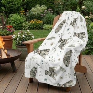 May include: A white throw blanket with a repeating pattern of gray dog illustrations, ferns, and floral accents, draped over a wooden chair. The blanket is displayed outdoors, suggesting a cozy, decorative item for home or garden use.
