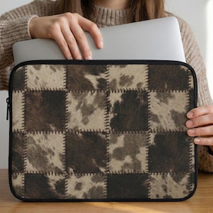 May include: Laptop sleeve with a patchwork design. The sleeve features a pattern of squares in shades of brown and tan, resembling a cowhide texture. The squares are stitched together with visible thread, and the sleeve has a black zipper.