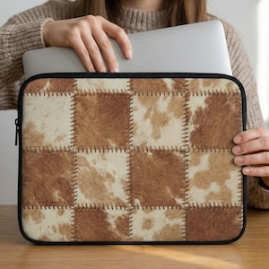 Puede incluir: Una funda para portátil con diseño de patchwork. La funda presenta cuadrados de piel sintética marrón y crema, cosidos con hilo marrón. La funda tiene una cremallera negra y un borde negro.