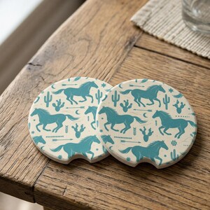 May include: Two round car coasters with a cream background and a teal horse and desert-themed pattern. The coasters are made of absorbent stone and have a small notch for easy removal from the cup holder.