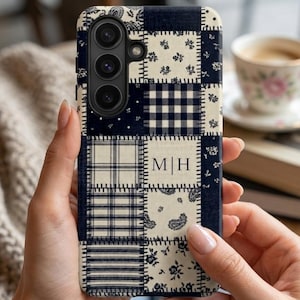 May include: A navy blue and cream patchwork phone case. The case has a variety of patterns, including floral, plaid, and paisley. The initials "M|H" are displayed in a rectangular block. The case is held in a person's hands.