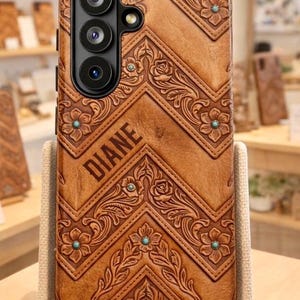 Könnte beinhalten: Eine braune Leder-Handyhülle mit floralem und geometrischem Design. Die Hülle trägt den Namen "DIANE" und türkisfarbene Akzente. Die Hülle wird auf einem neutralfarbenen Ständer präsentiert.