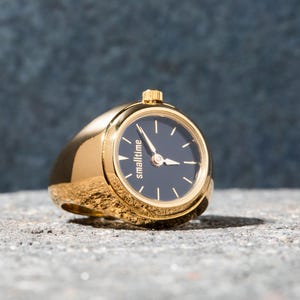 Peut inclure: Une montre-bague dorée avec un cadran bleu foncé et des aiguilles dorées. Le cadran de la montre porte l'inscription "smalltime". La bague repose sur une surface grise texturée.