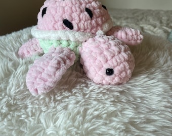 May include: A handmade plush turtle in the shape of a watermelon. The turtle has a pink body with black seed details on the shell. The underside of the shell is green, and the edges are white. The turtle is lying on a white, fluffy surface.