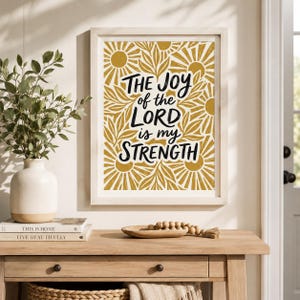 May include: A framed wall art print featuring the black text THE JOY of the LORD is my STRENGTH centered over a mustard yellow and white pattern of stylized suns and leaves. The print is displayed in a light-colored wooden frame.