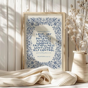 May include: Framed print with a cream background and blue floral border. The text reads "Love, Joy, Peace, Patience, Kindness, Goodness, Faithfulness, Gentleness, Self Control" from Galatians 5:22-23. A vase with dried flowers sits to the right.
