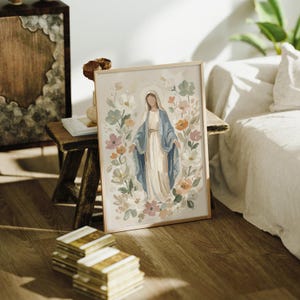 May include: Framed art print featuring a watercolor painting of a religious figure in a blue robe and white dress, surrounded by pastel-colored flowers. The artwork has a light beige background and is displayed in a wooden frame.
