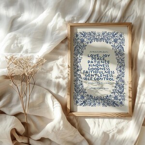 May include: A framed print with a light wood frame. The print features a floral border in blue surrounding a list of words: Love, Joy, Peace, Patience, Kindness, Goodness, Faithfulness, Gentleness, Self Control. The text is from Galatians 5:22-23.