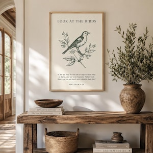 May include: A framed wall art print featuring a black line drawing of a bird perched on a branch with the text LOOK AT THE BIRDS and the scripture Matthew 6:26 printed in black on a cream background, displayed in a light wood frame.
