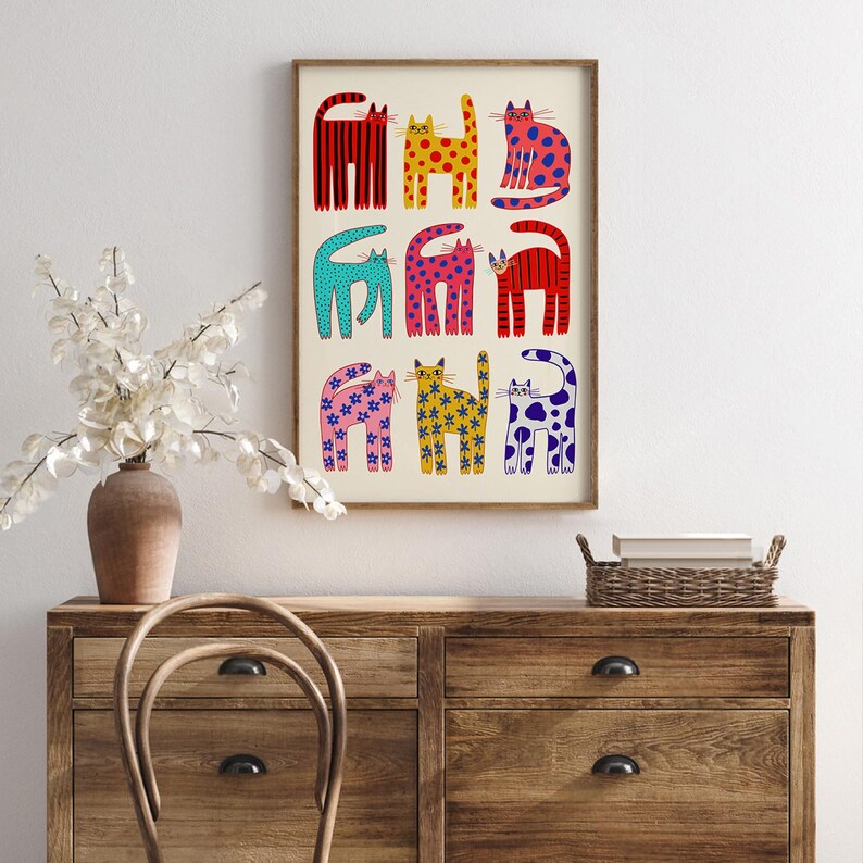 May include: A framed art print featuring nine colorful cat illustrations. Each cat is uniquely designed with patterns and colors, including red stripes, yellow spots, and blue polka dots. The artwork is displayed on a cream-colored background within a wooden frame.