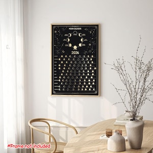 May include: A framed black and gold moon calendar for the year 2026. The calendar displays the phases of the moon and constellations. The text "Moon Calendar" and "2026" are visible. The frame is a light brown color.
